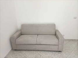 Un divano semplice e chiaro in un angolo della stanza, con pavimento piastrellato e parete bianca liscia.