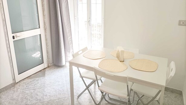 A room with a white table, four chairs, a window with curtains, and a door with a frosted glass panel. - Newhouse Class (Bari)