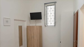 A room with a wooden wardrobe, a television mounted on the wall, a window with bars, and a door. - Newhouse Class (Bari)