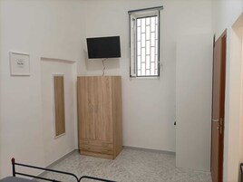 Una stanza con un armadio in legno, una televisione fissata al muro, una finestra con le grate e una porta.