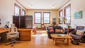 A spacious living room with a wooden desk, leather chairs, a sofa, and a coffee table. There are large windows with blinds, a bookshelf, and a painting on the wall. - Historic High School - Stays and events close to skiing, golf, fishing, hunting! (Kingston)