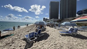 A beach with sun loungers and umbrellas, a clear sky, and tall buildings in the background. - Exclusive 3 Bedrooms Apartment W/ Amazing View (Hallandale Beach)