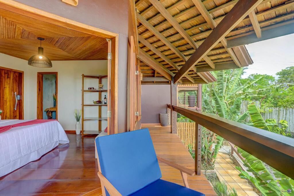 A room with a bed, a blue chair, wooden flooring, a wooden ceiling, and a balcony with a view of greenery.