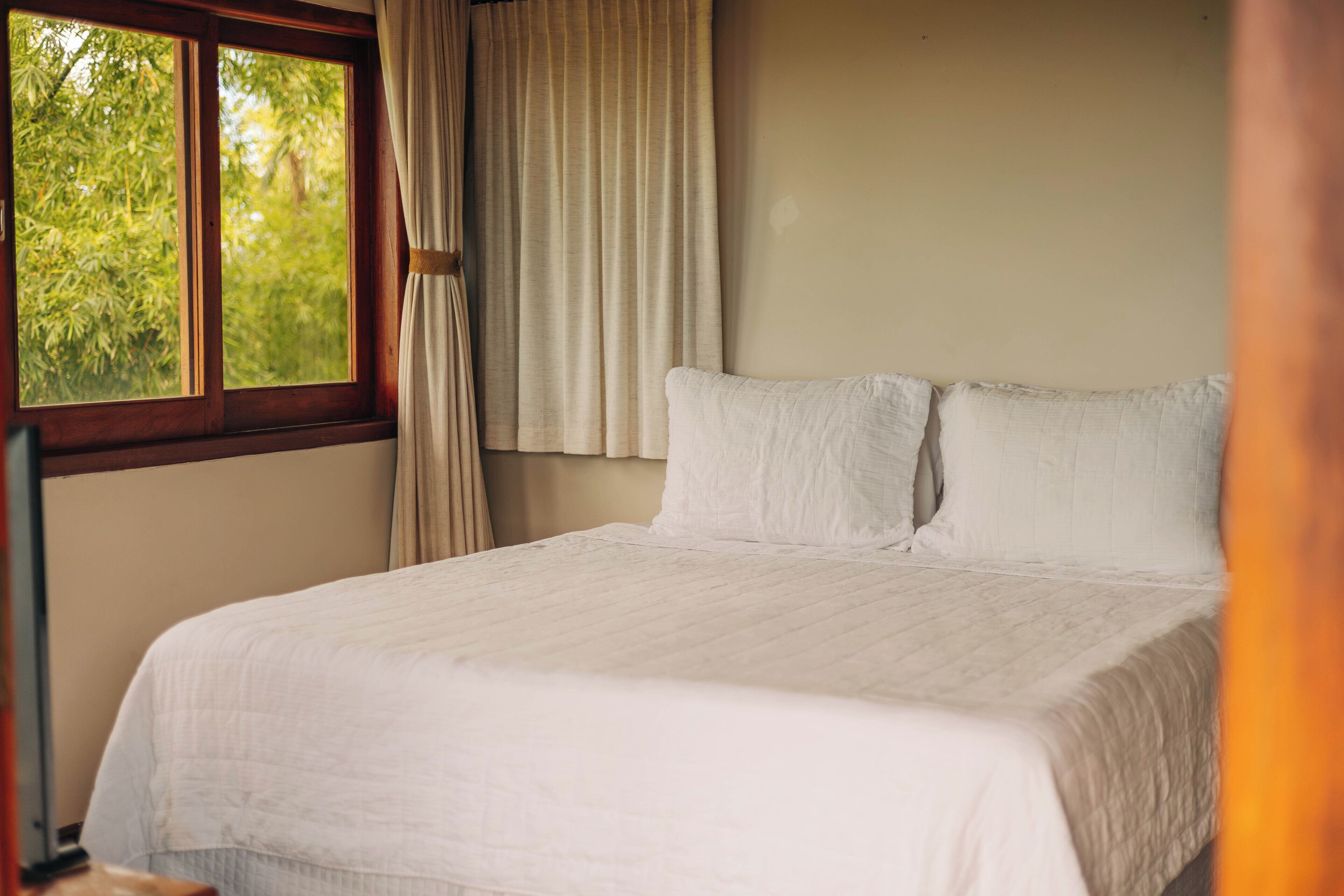 Una cama bien hecha con sábanas blancas en una habitación con un ventanal y cortinas.