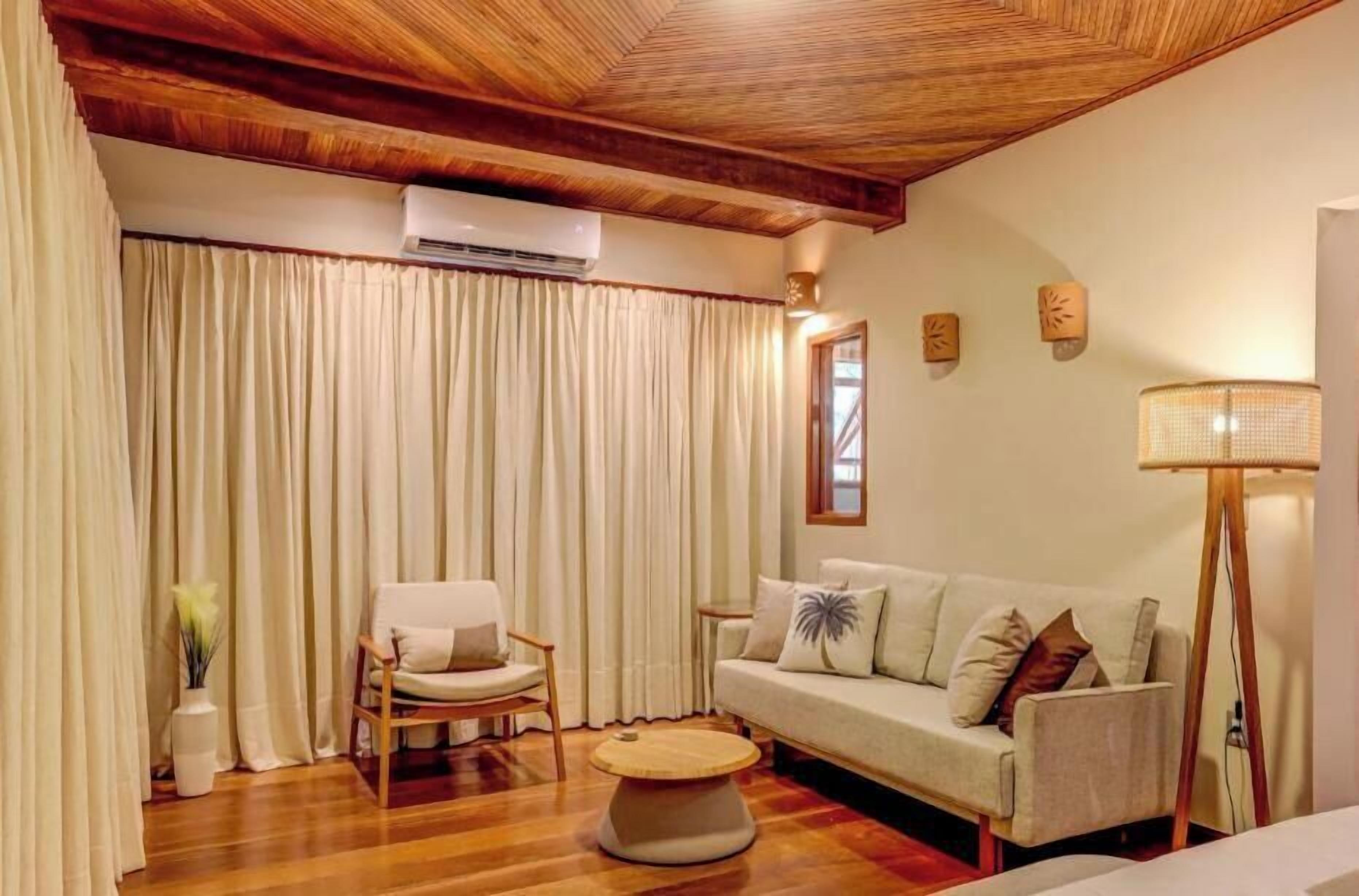 A room with a wooden ceiling, a sofa, a chair, a small round table, and a floor lamp.
