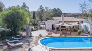 A poolside area with lounge chairs, a covered patio, and a building with a tiled roof. - Domu Prexada (Maracalagonis)