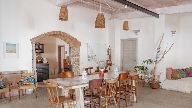 A spacious dining area with a large wooden table, multiple chairs, hanging woven light fixtures, and a stone pillar. - Domu Prexada (Maracalagonis)