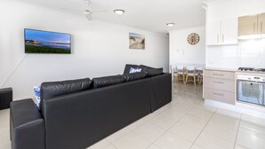 A modern living room with a black sofa, a flat-screen TV, and a dining area with a white table and chairs. - Stylish Unit with Water Views (BELLARA)