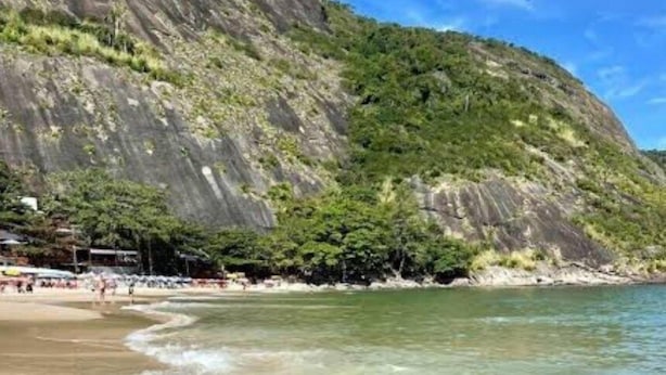 A beach with clear shallow waters, a rocky cliff, and a few people in the distance. - Andorinhas (Niteroi)