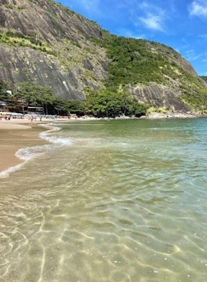 A beach with clear shallow waters, a rocky cliff, and a few people in the distance. - Andorinhas (Niteroi)