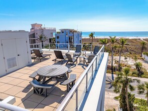 Terraço no telhado com mesas e cadeiras, com vista para uma praia e palmeiras.