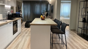 Een moderne keuken met een centraal eiland, een eetgedeelte met stoelen en een zicht op een slaapkamer door een deuropening.