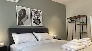 Een netjes opgemaakt bed met witte lakens, een nachtkastje met een lamp, en twee ingelijste botanische prints aan de muur.