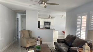 Una sala de estar con un sofá de cuero marrón, un sillón beige y una mesa de centro de madera. Se aprecia una cocina al fondo, con armarios blancos y un microondas negro.
