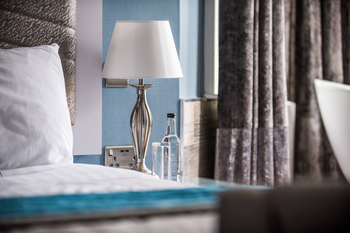 A hotel room with a bed, a bedside table with a lamp, a bottle, and a power outlet.