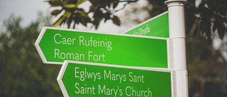 A directional sign with green panels indicating directions to Caer Rufeinig Roman Fort and Eglwys Marys Sant Saint Mary's Church.