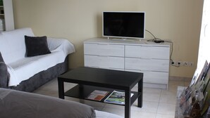 Una habitación con un sofá blanco, una mesa de centro negra, un aparador blanco y un televisor encima.
