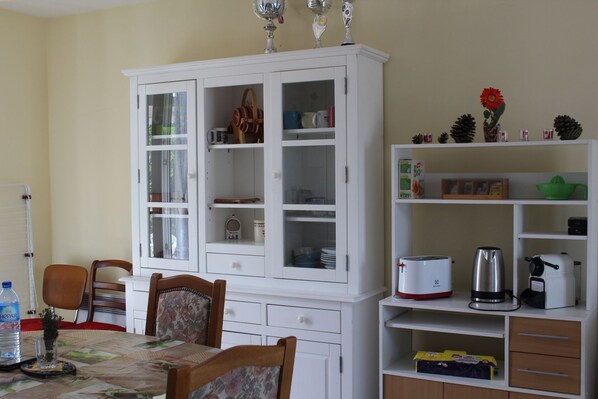 A dining area with a white cabinet, chairs, a table with a placemat, and various kitchen appliances.