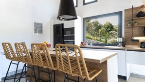 A modern kitchen with a central island, bar stools, and a large window overlooking greenery.
