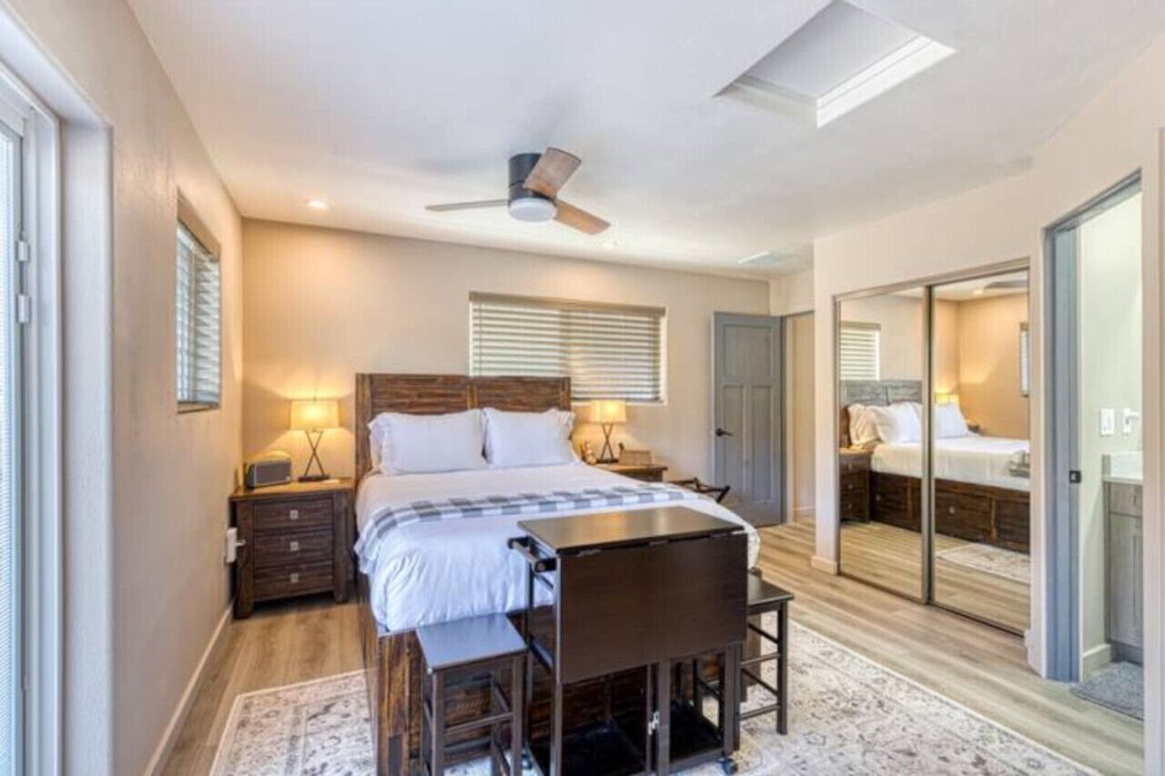A bedroom with a bed, bedside tables, a desk, and a chair. There is a ceiling fan, a window with blinds, and a sliding glass door leading to another room.