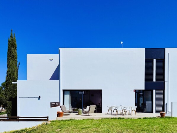 Una casa moderna de dos pisos con techo plano, ventanas amplias y un patio con mobiliario exterior.