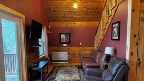 A cozy cabin interior with a wooden ceiling, a fireplace, a television, and a large window.