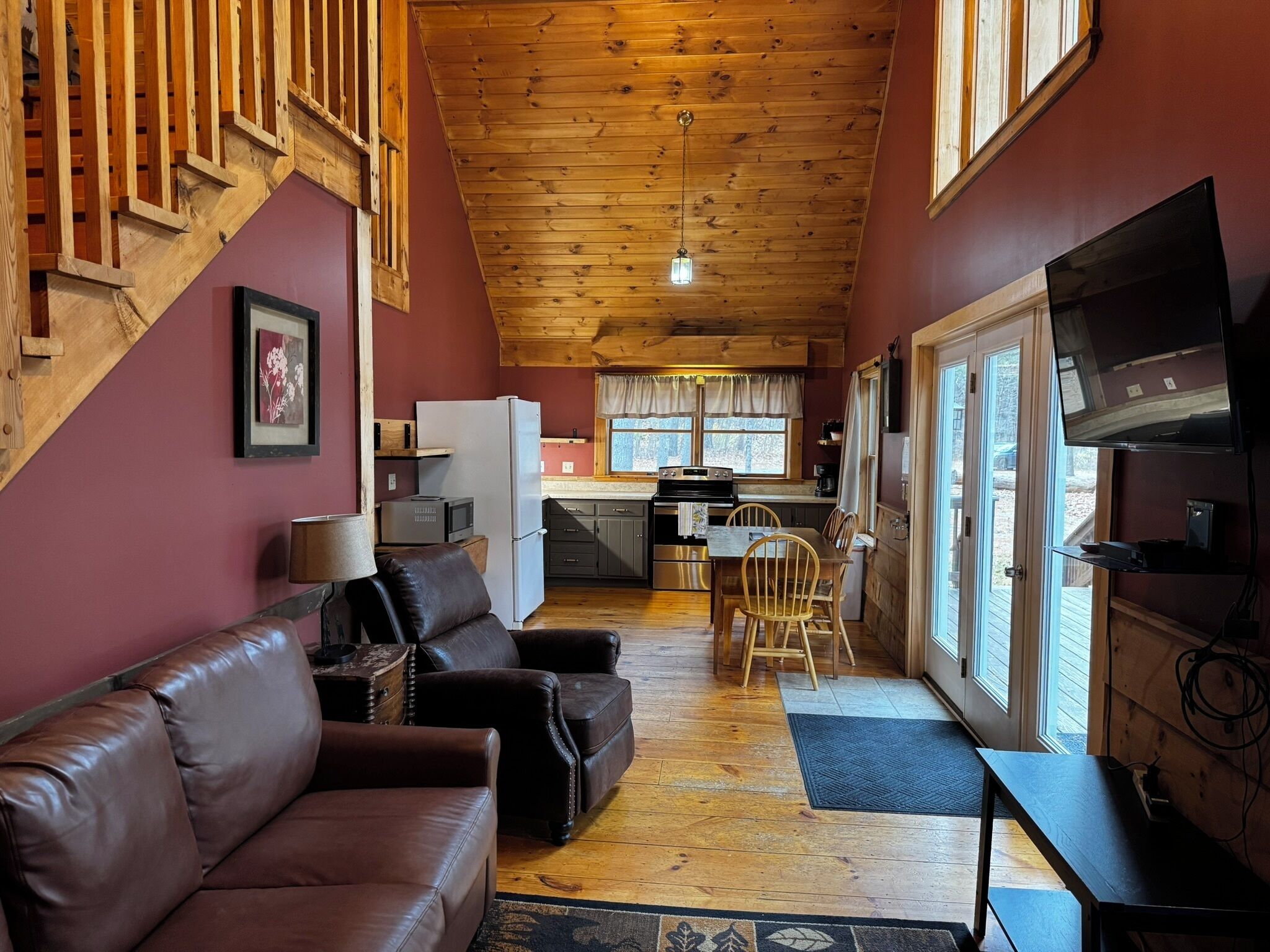 A cozy cabin interior with a wooden ceiling, a living area with a sofa and a TV, a kitchen with a refrigerator and stove, and a dining area with chairs.