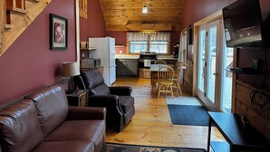 A cozy cabin interior with a wooden ceiling, a living area with a sofa and a TV, a kitchen with a refrigerator and stove, and a dining area with chairs.