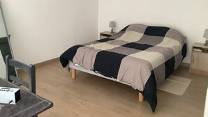 Un lit simple avec une couvre-lit à carreaux, dans une pièce dotée d’un plancher en bois et de murs blancs. On y trouve une table de chevet avec une lampe, ainsi qu’une petite table sur laquelle est posée une boîte.