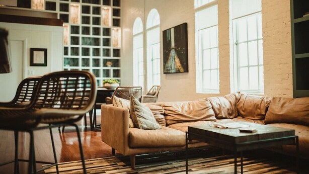 A spacious living room with a large sofa, a coffee table, and a wicker chair. There are large windows with arched tops, a brick wall, and a framed painting on the wall. - 5-star Luxury Apartment - Atop the Seagle Building (Gainesville)