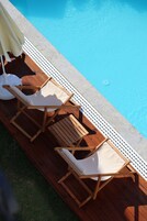 Uma área à beira da piscina com espreguiçadeiras de madeira e um guarda-sol branco.