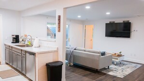 A modern kitchen with white cabinets, a coffee maker, and a trash bin. A living area with a sofa and a TV is visible through an open door. - Updated Ann Arbor ranch w/private yard and lots of parking!  (Ann Arbor)