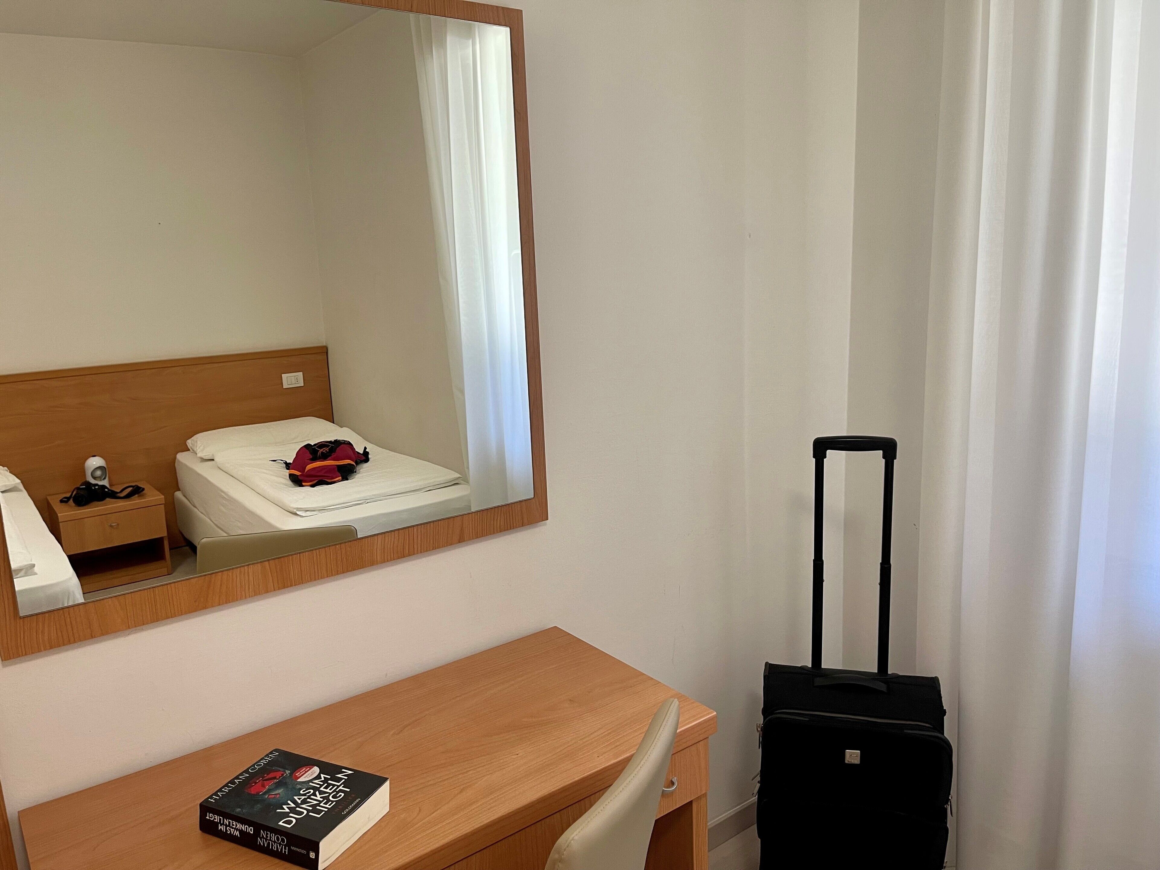 A hotel room with a wooden desk, a black suitcase, a book, and a bed with a white comforter.