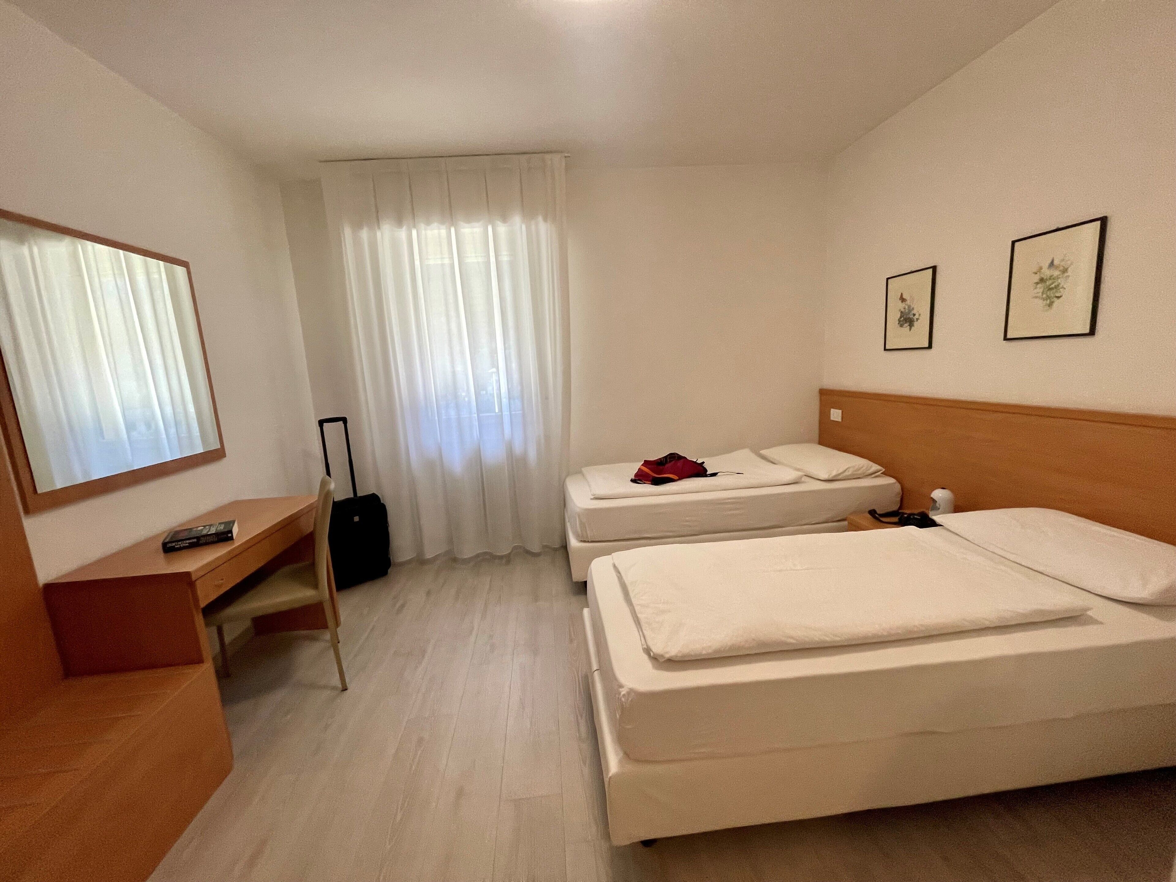 A hotel room with two beds, a desk, a chair, a suitcase, and a mirror.