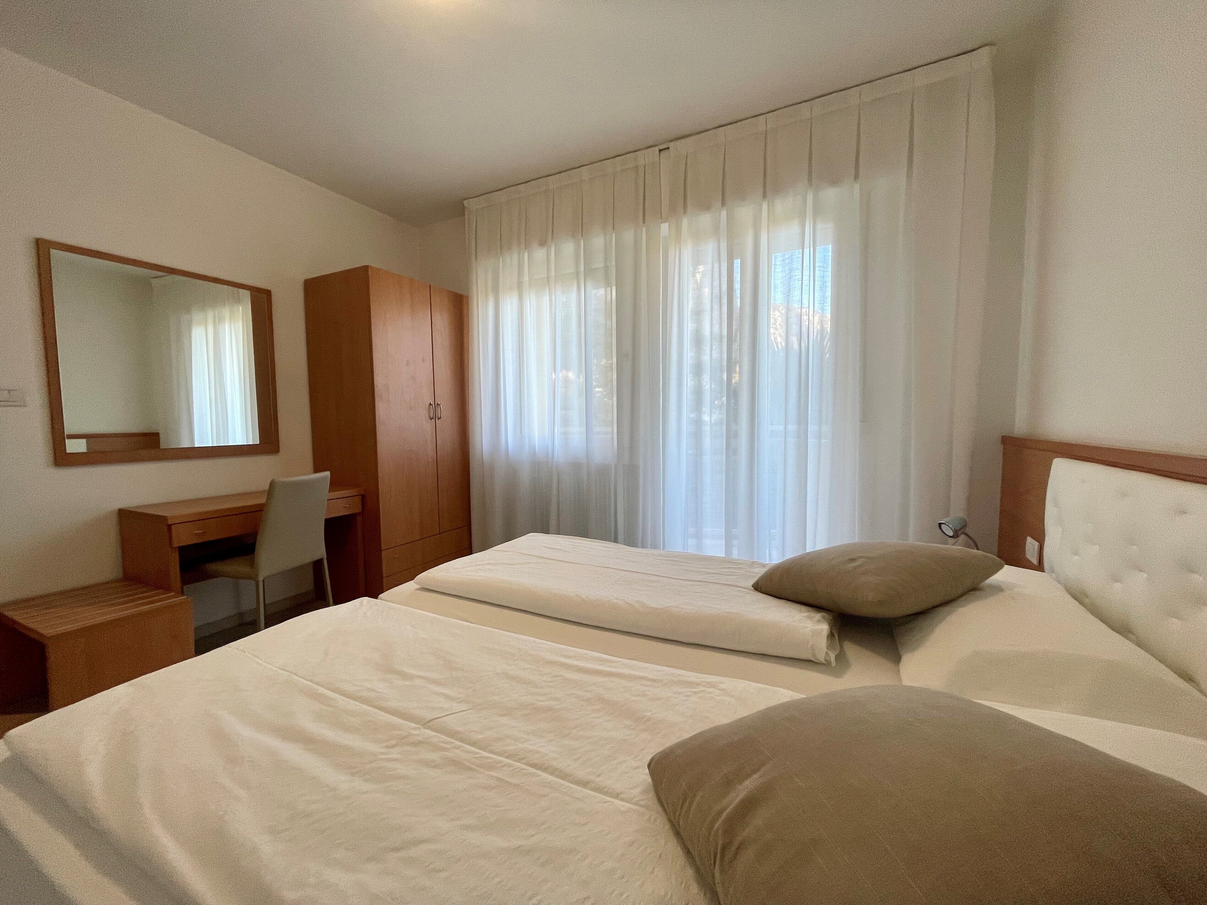 A hotel room with a bed, a desk, a chair, a wardrobe, and a large window with curtains.
