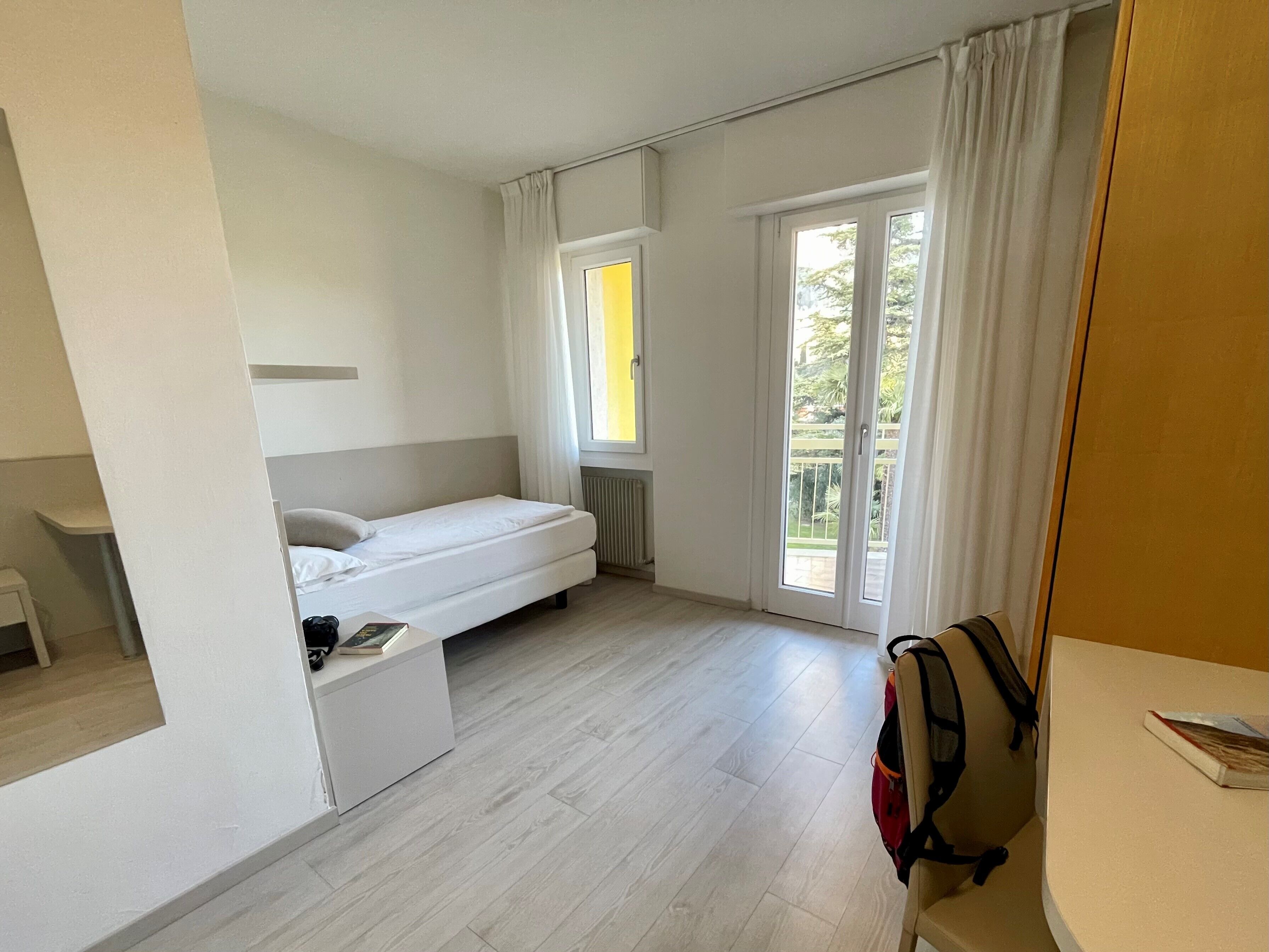 Ett kompakt hotellrum med en enkel säng, ett litet skrivbord och en skjutdörr i glas som leder ut till en balkong.