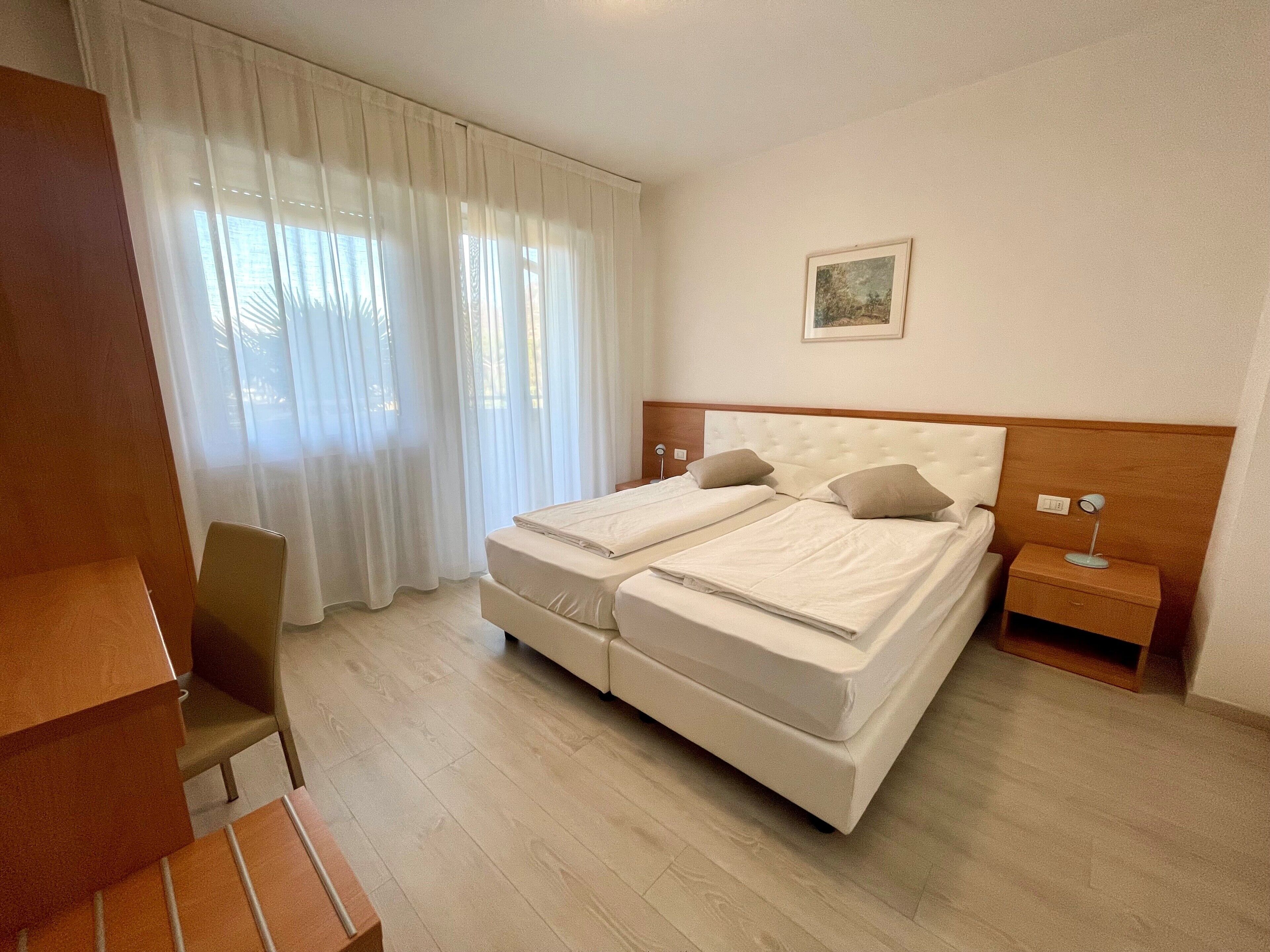 Quarto de hotel com cama de casal, uma cadeira, mesa de cabeceira e janela com cortinas leves.