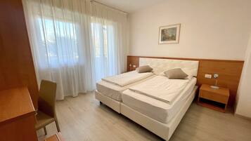 Quarto de hotel com cama de casal, uma cadeira, mesa de cabeceira e janela com cortinas leves.