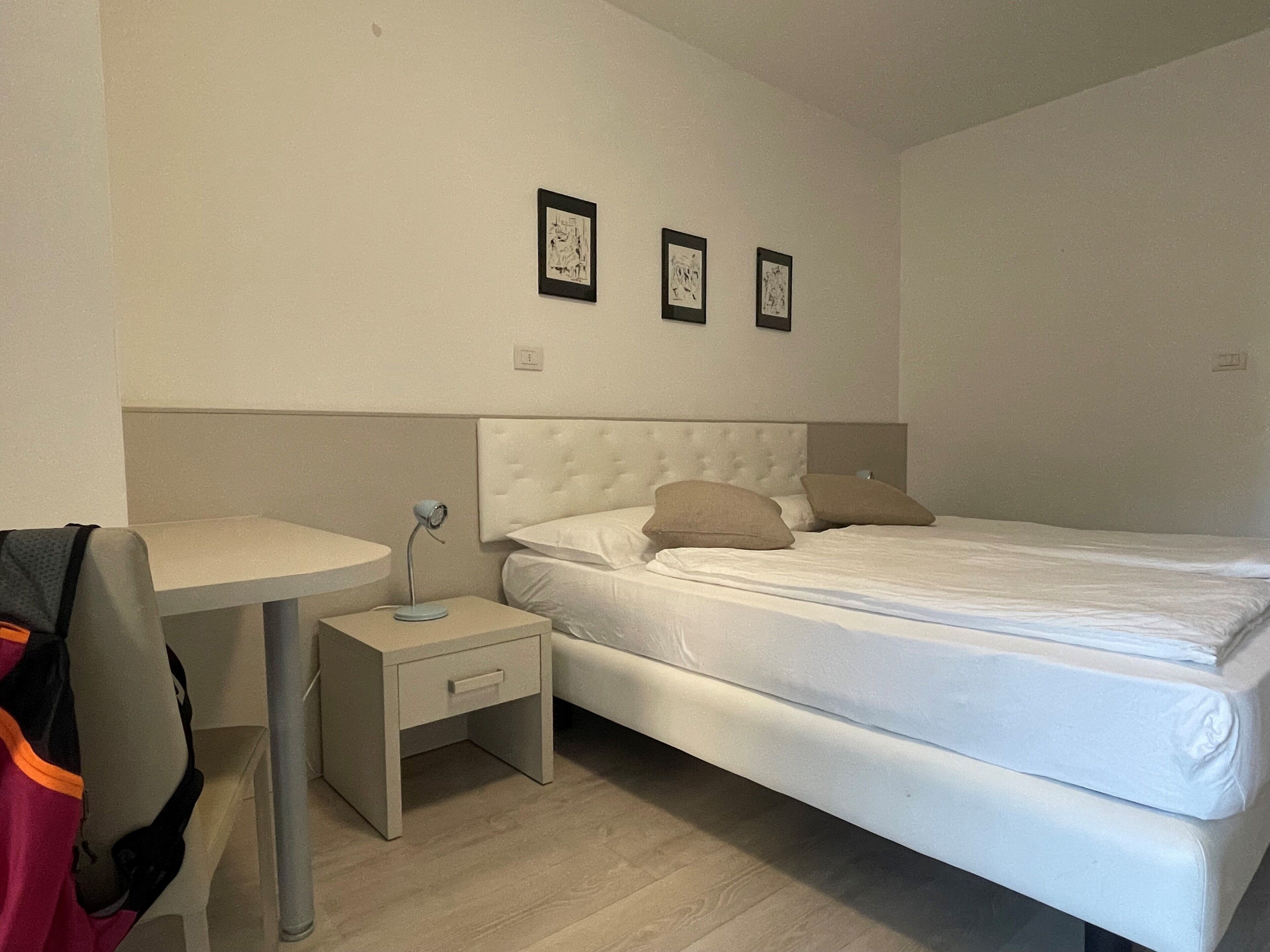 A bedroom with a bed, a bedside table, and a small desk. There are three framed pictures on the wall above the bed.
