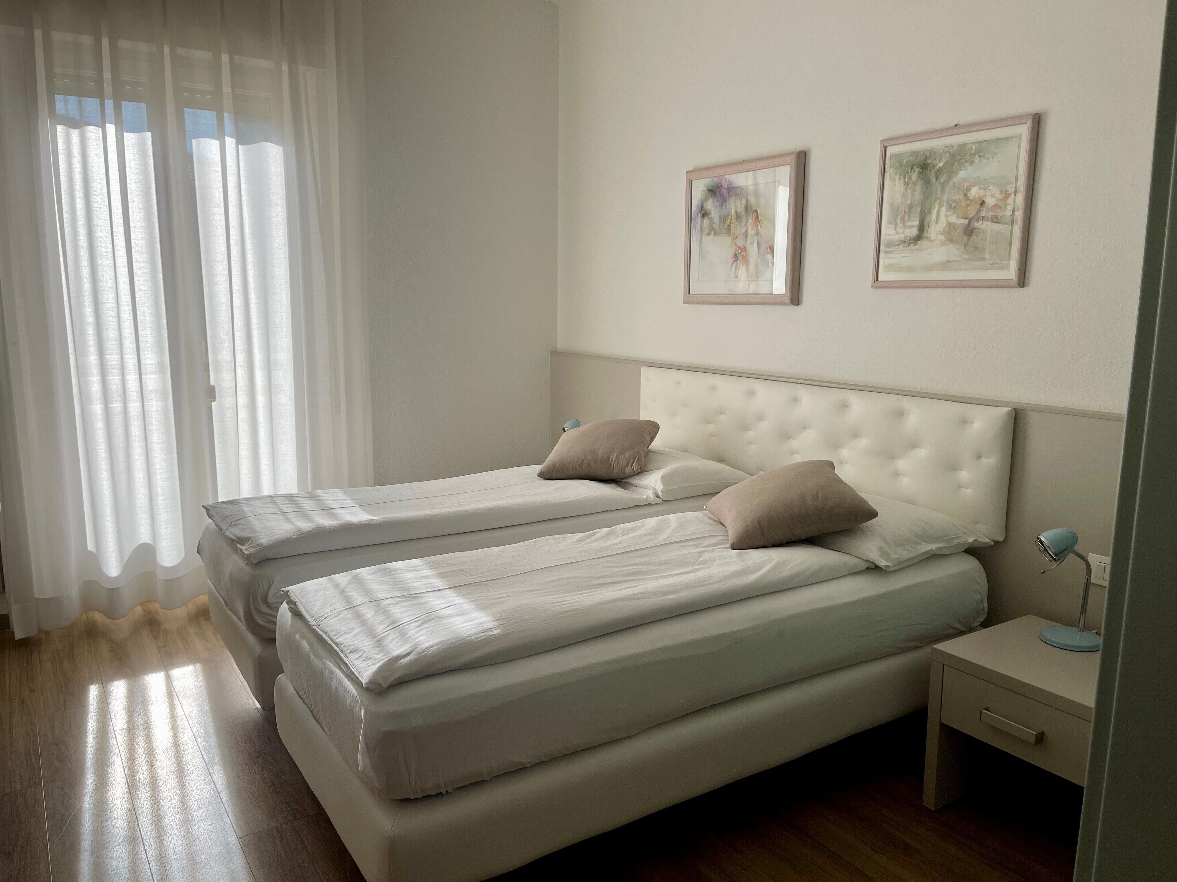 Un dormitorio con dos camas, una mesita de noche y una ventana con cortinas traslúcidas.