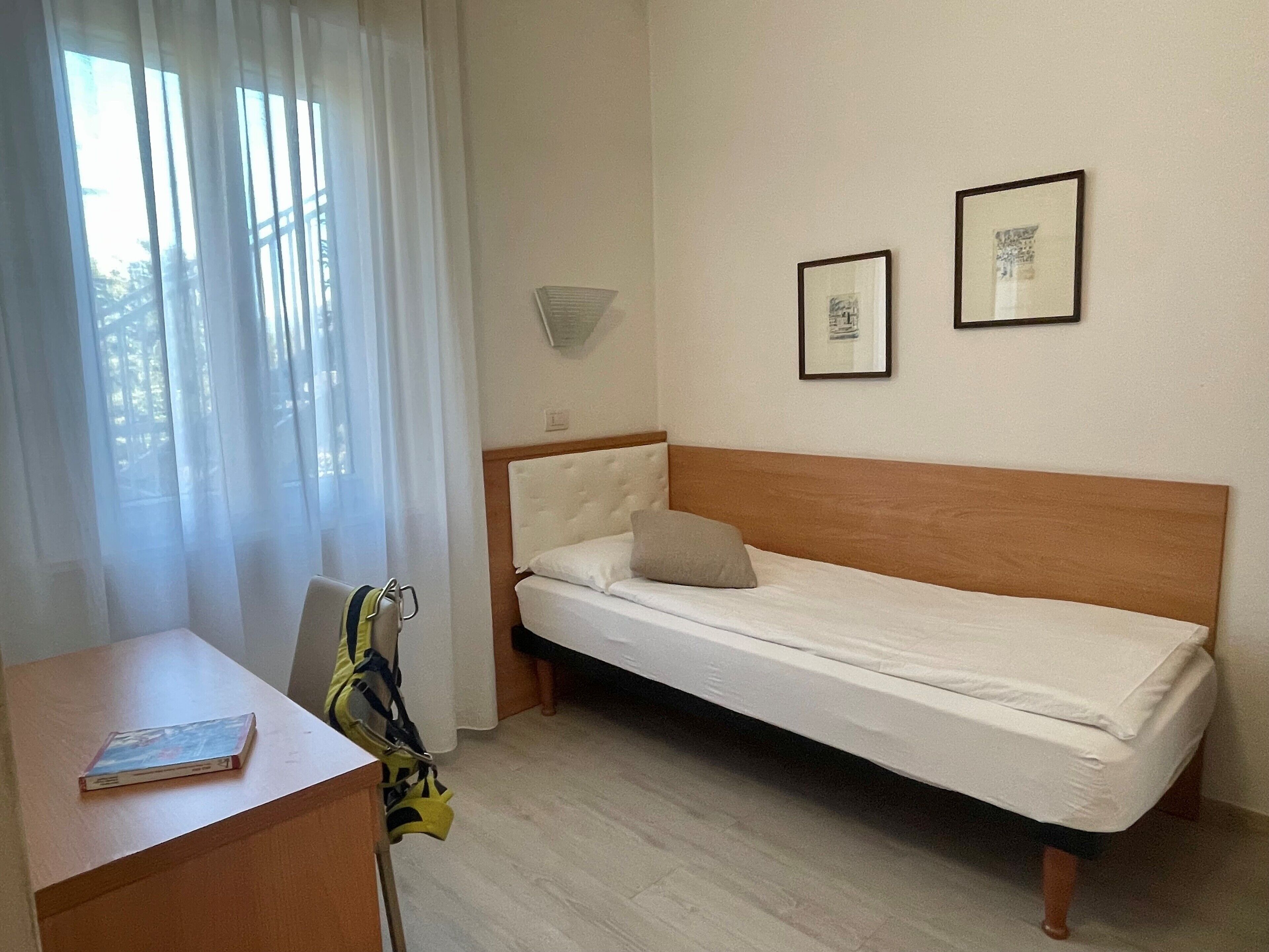 Quarto pequeno com uma cama de solteiro, cabeceira de madeira, escrivaninha com cadeira e duas imagens emolduradas na parede.