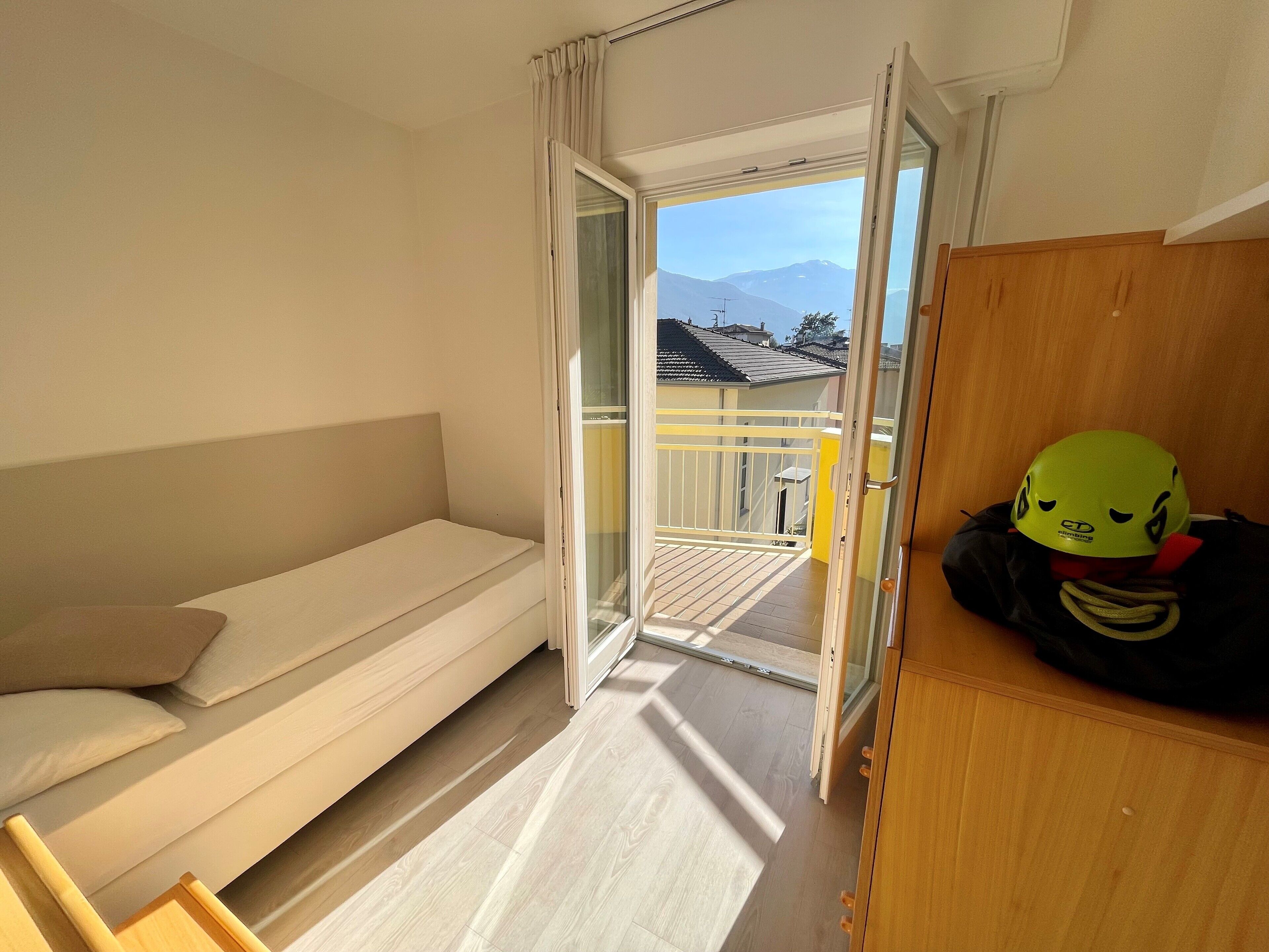 Una habitación con una cama, un armario de madera y un balcón con vistas a casas y montañas.