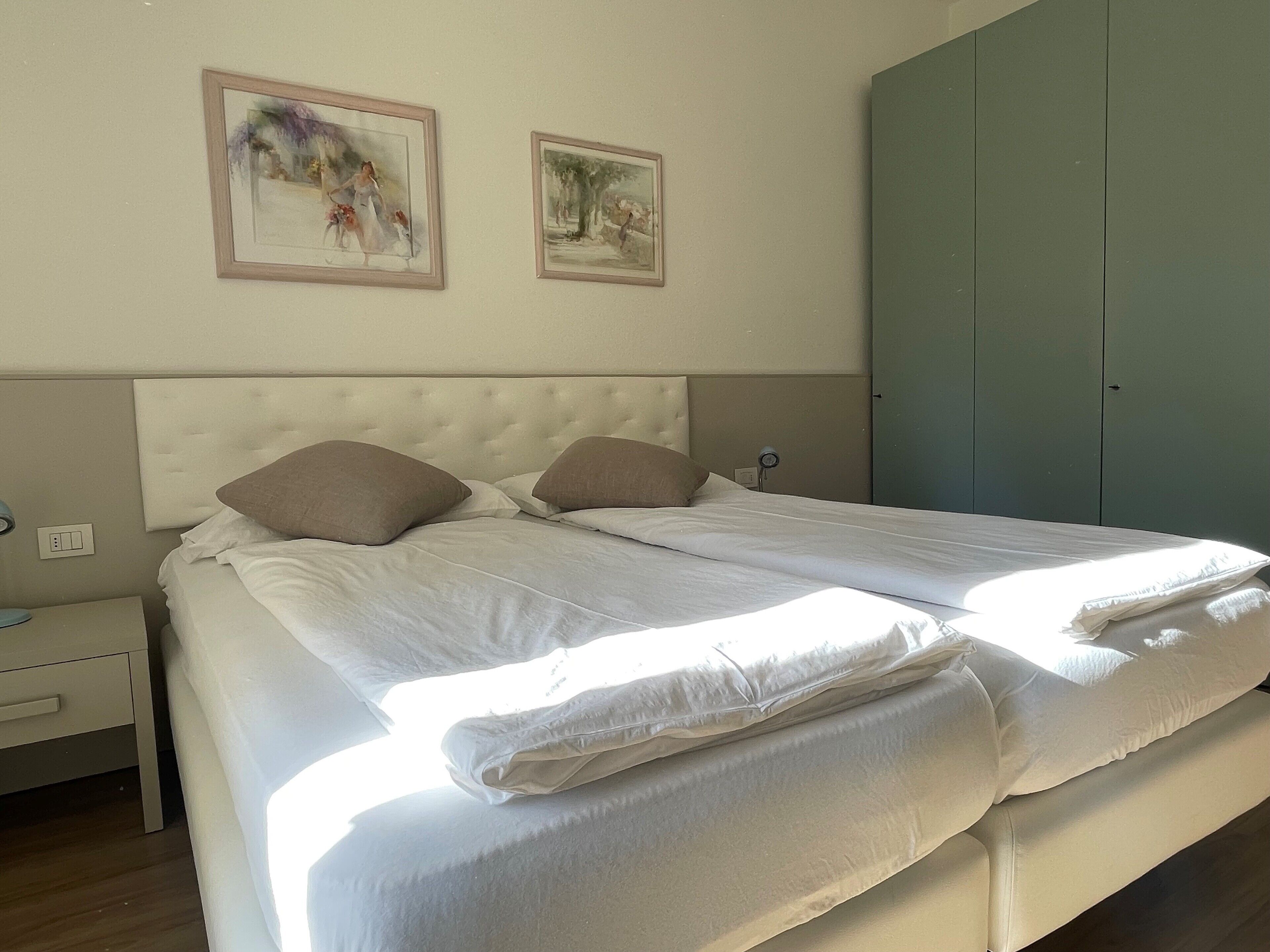 Una cama doble con ropa blanca, un cabecero capitonado y dos cuadros enmarcados en la pared.