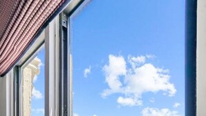 A window with sheer white curtains, a clear blue sky, and a parking lot with cars and buildings in the background.