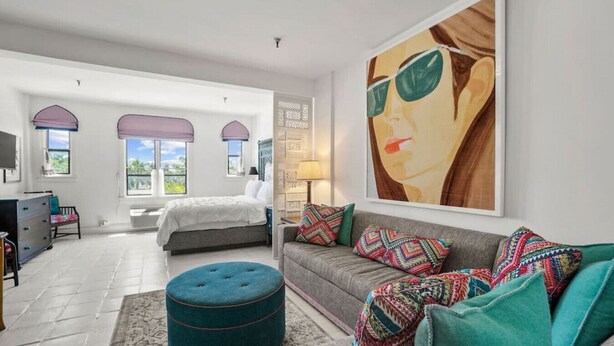 A spacious bedroom with a large bed, a grey sofa, colorful cushions, a teal ottoman, and a large painting on the wall.