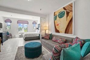 Un dormitorio amplio con una cama grande, un sofá gris, cojines de colores, un otomano color turquesa y un cuadro grande en la pared.