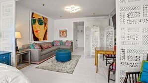 A modern living room with a grey sofa, colorful cushions, a blue ottoman, a wooden coffee table, and a large abstract painting.