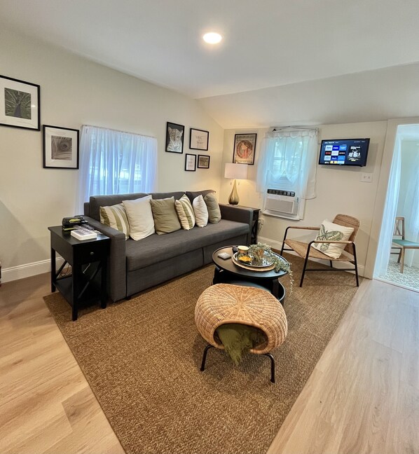 Un salon comprenant un canapé, une table basse, un pouf en osier, un téléviseur fixé au mur et une fenêtre avec des rideaux.