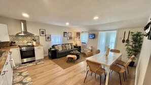 A modern kitchen with a stove, oven, and sink. There is a dining area with a table and chairs. The room has a sofa and a TV on the wall.