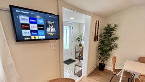 A living room with a flat-screen TV displaying various streaming services, a wicker chair with a butterfly pillow, a dining area with a table and chairs, and a potted plant.
